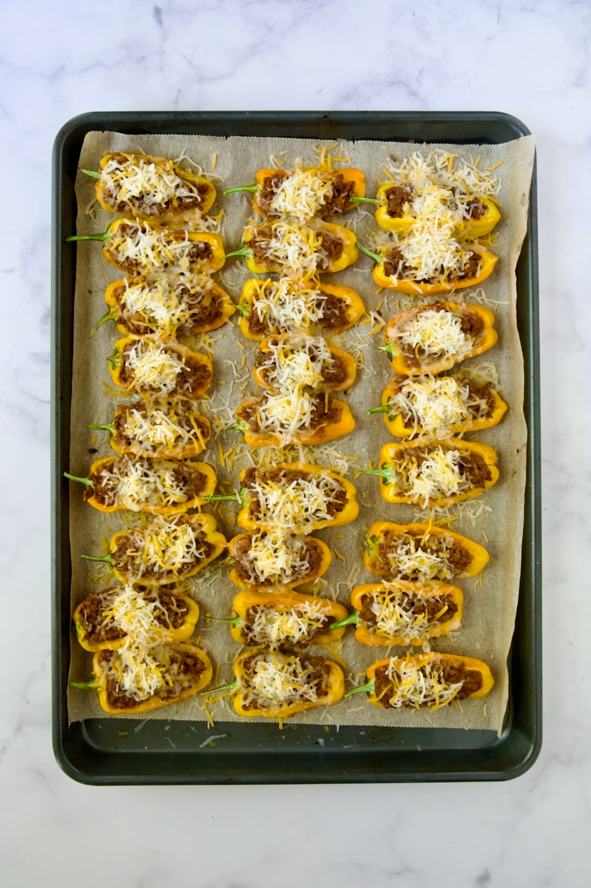 Halved, partially baked yellow mini peppers on parchment paper lined baking sheet with shredded Mexican cheese blend added.