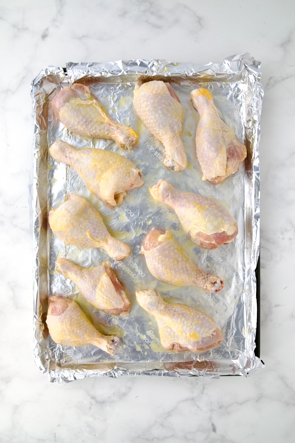 Ten uncooked unseasoned chicken drumsticks on aluminum foil-lined rimmed sheet pan.