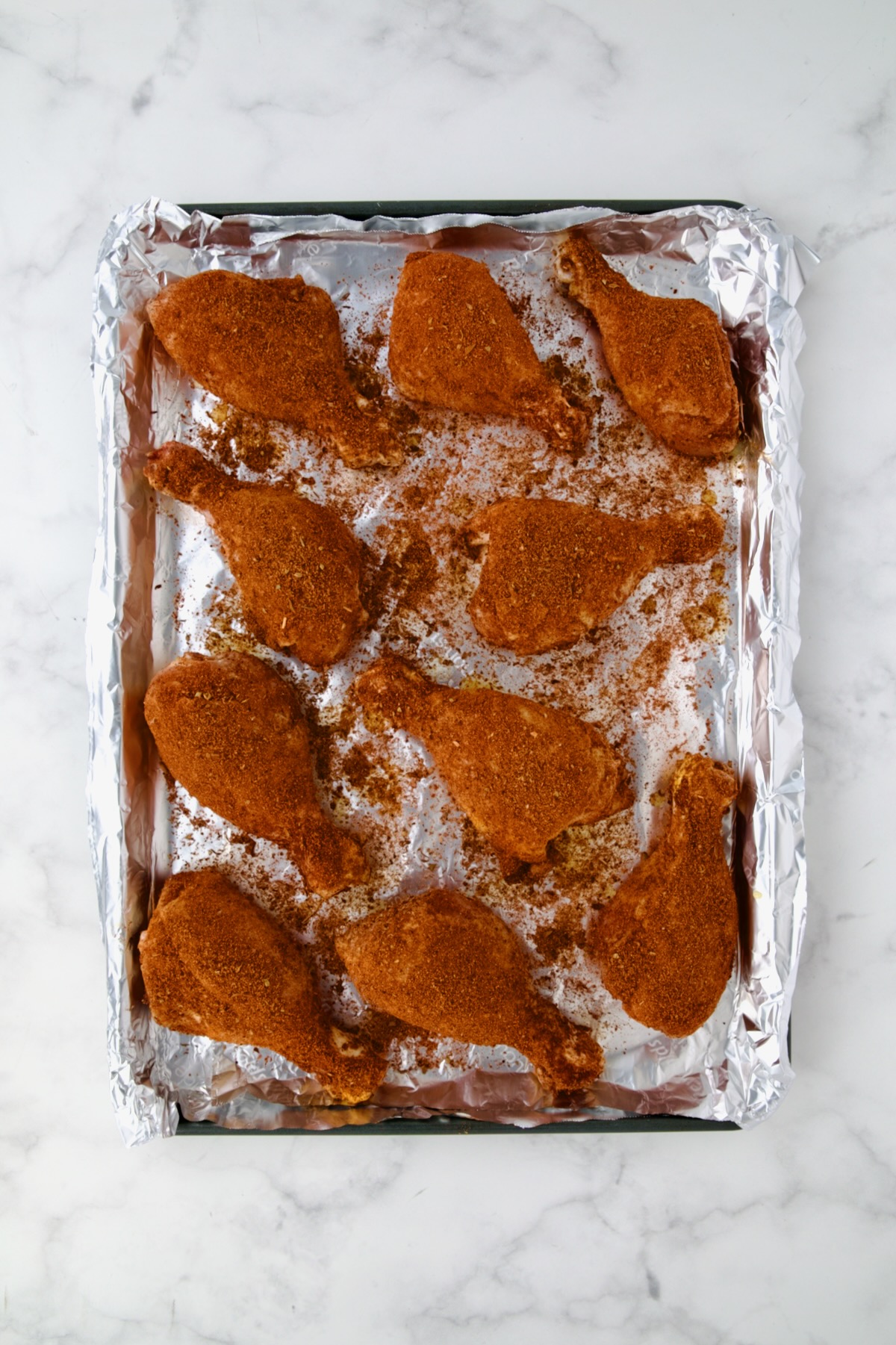 Ten uncooked seasoned chicken drumsticks on aluminum foil-lined rimmed sheet pan.