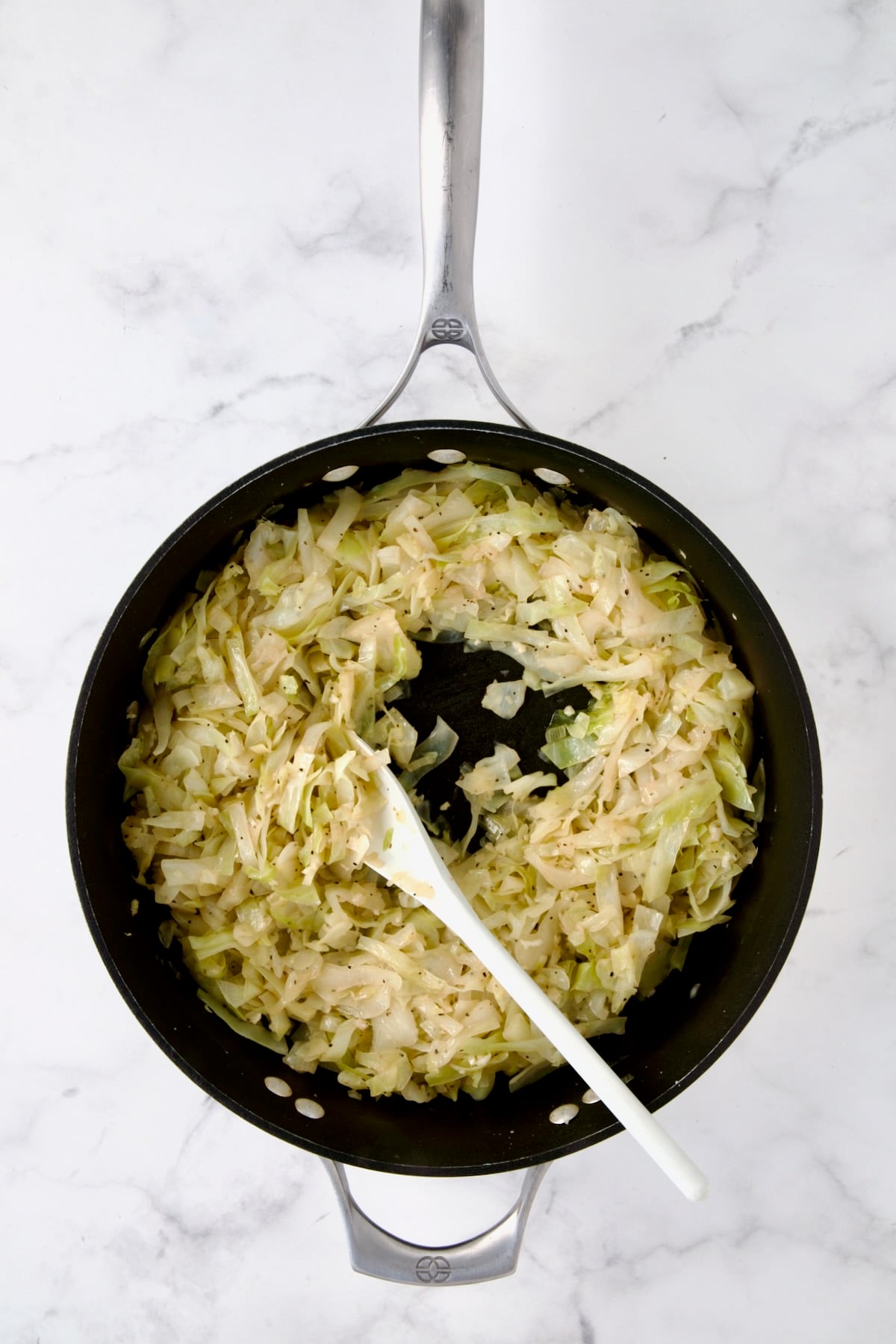 Cooked cabbage in nonstick skillet being stirred with white mixing spoon.