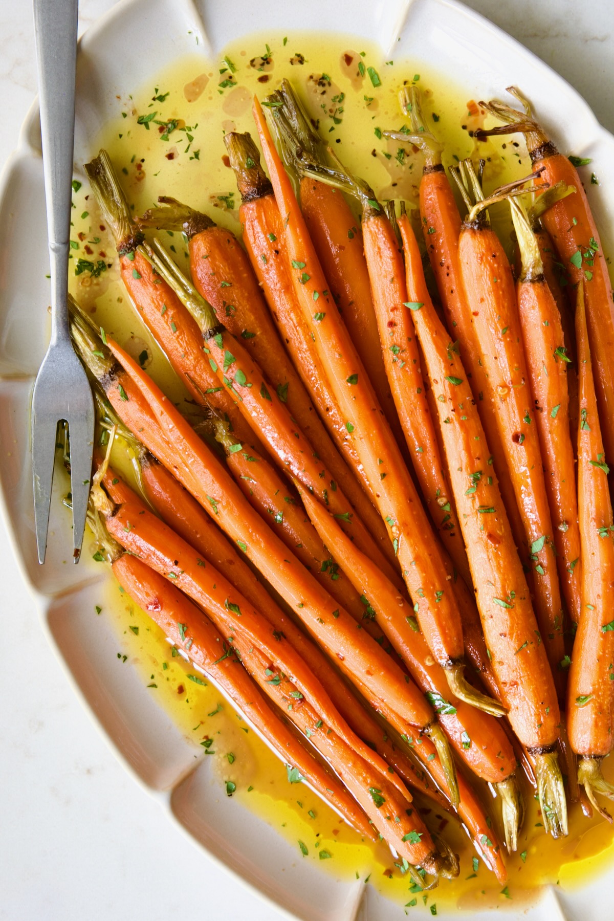 Hot Honey Roasted Carrots on white oval platter with serving fork garnished with fresh parsley.