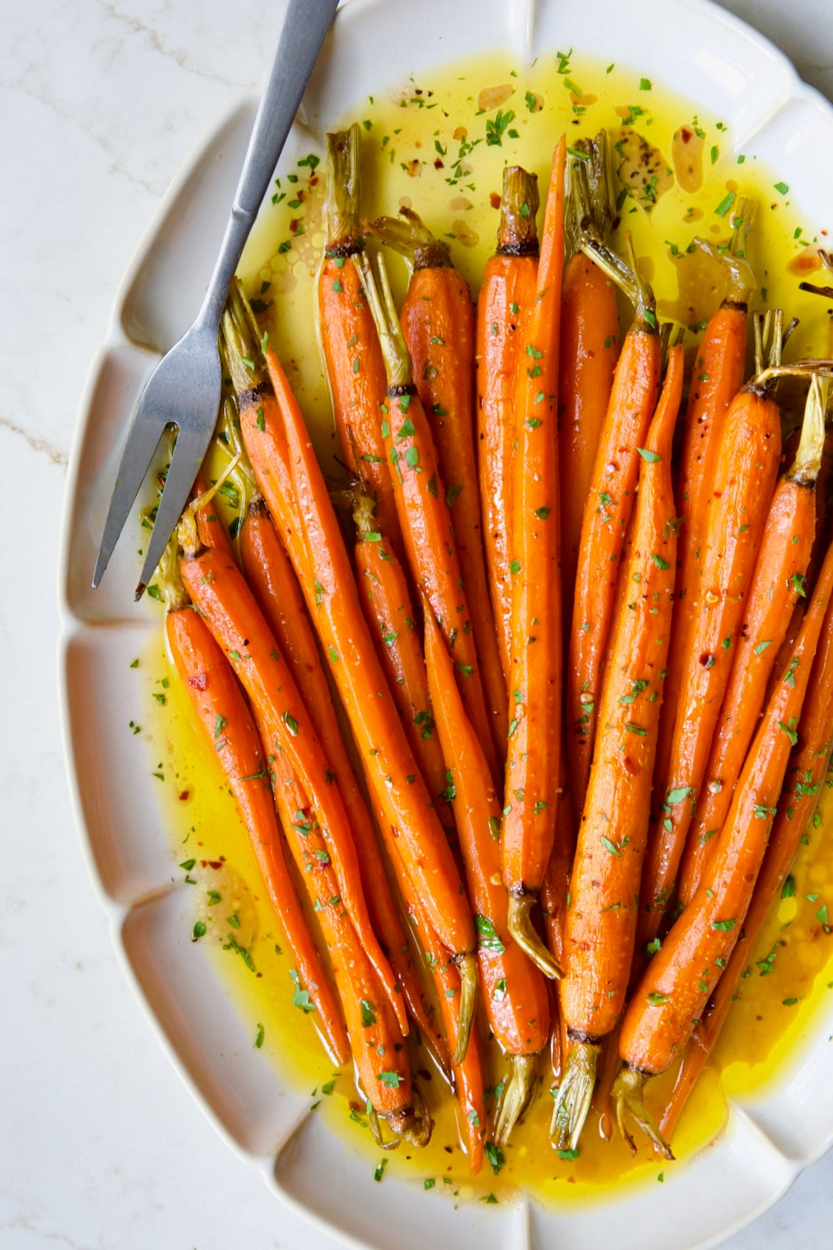 Hot Honey Roasted Carrots on white oval platter with serving fork garnished with fresh parsley.