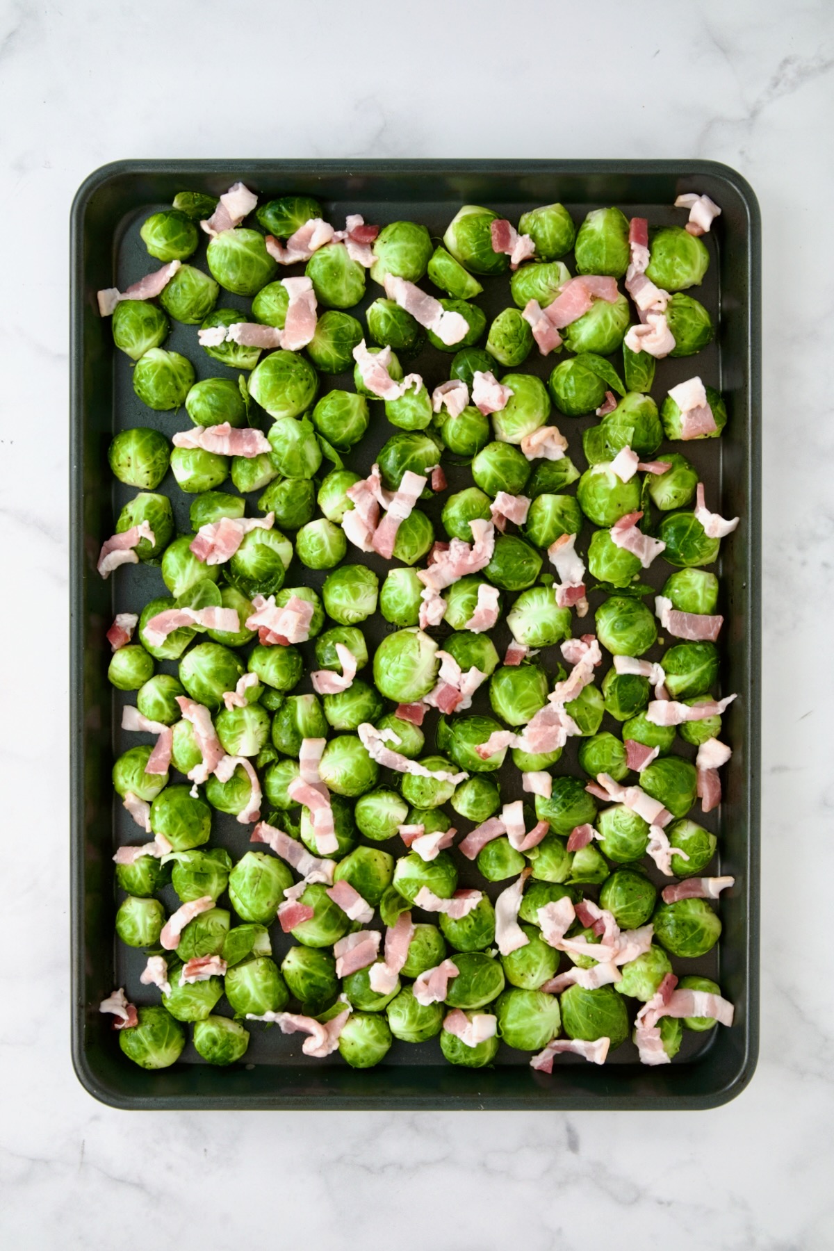 Halved Brussels sprouts on rimmed sheet pan with diced bacon.