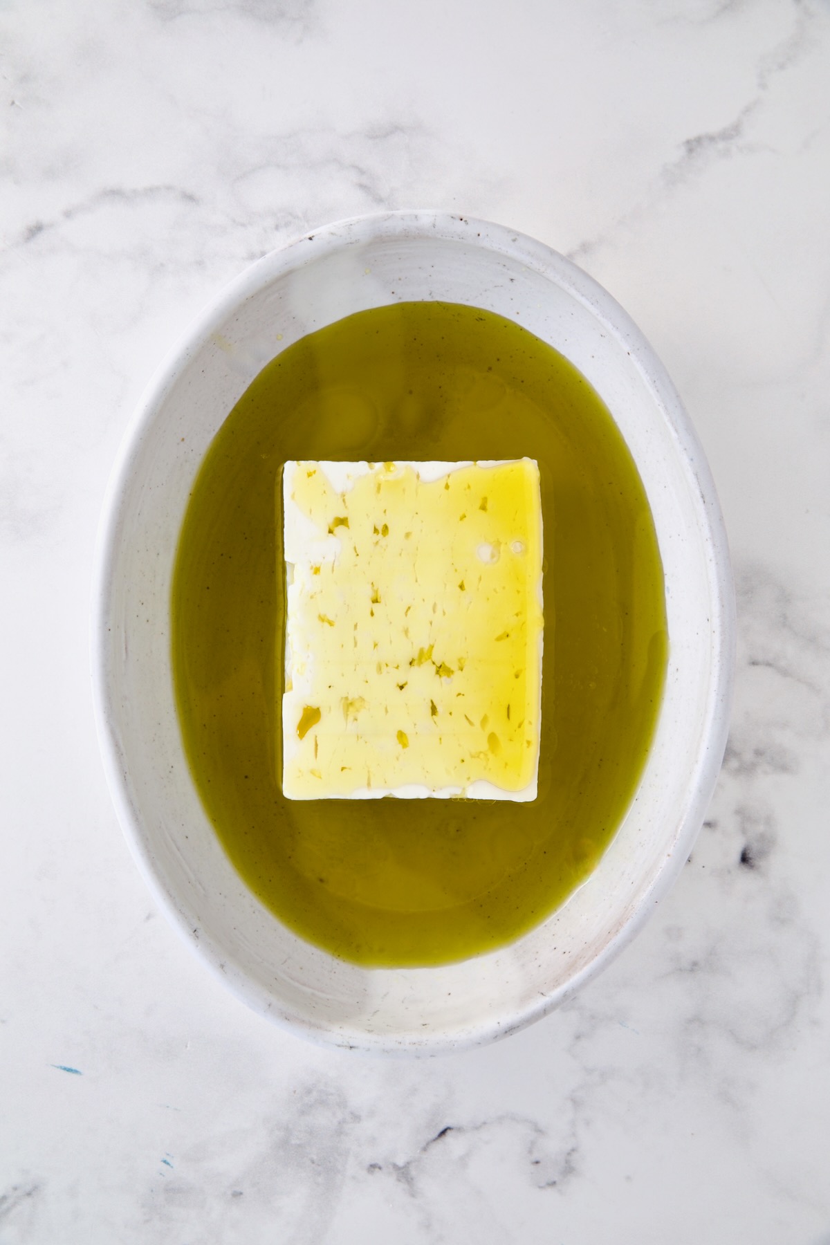 Block of feta cheese in oval white baking dish with olive oil poured over it.