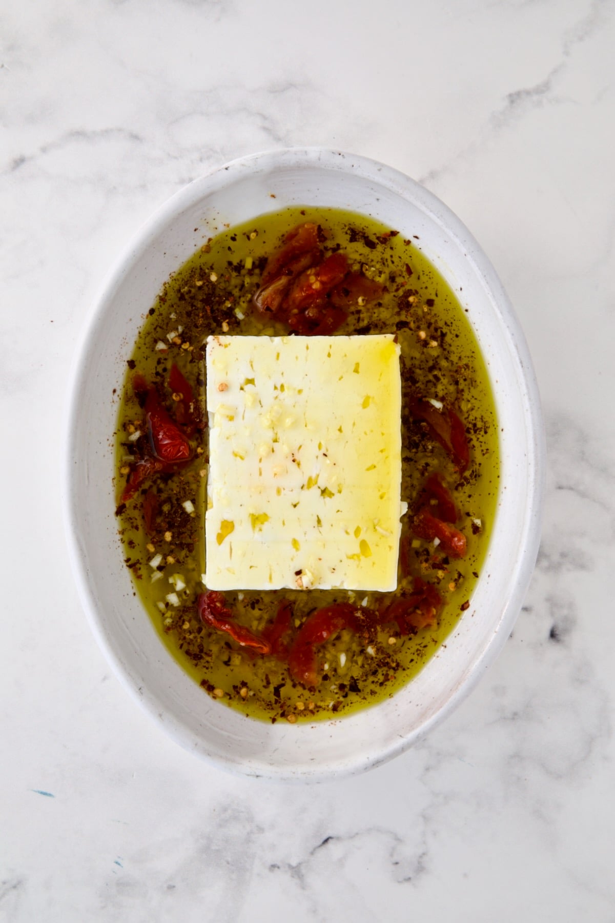 Block of feta cheese in oval white baking dish with olive oil poured over it and garlic, crushed red pepper flakes and sun-dried tomatoes added.
