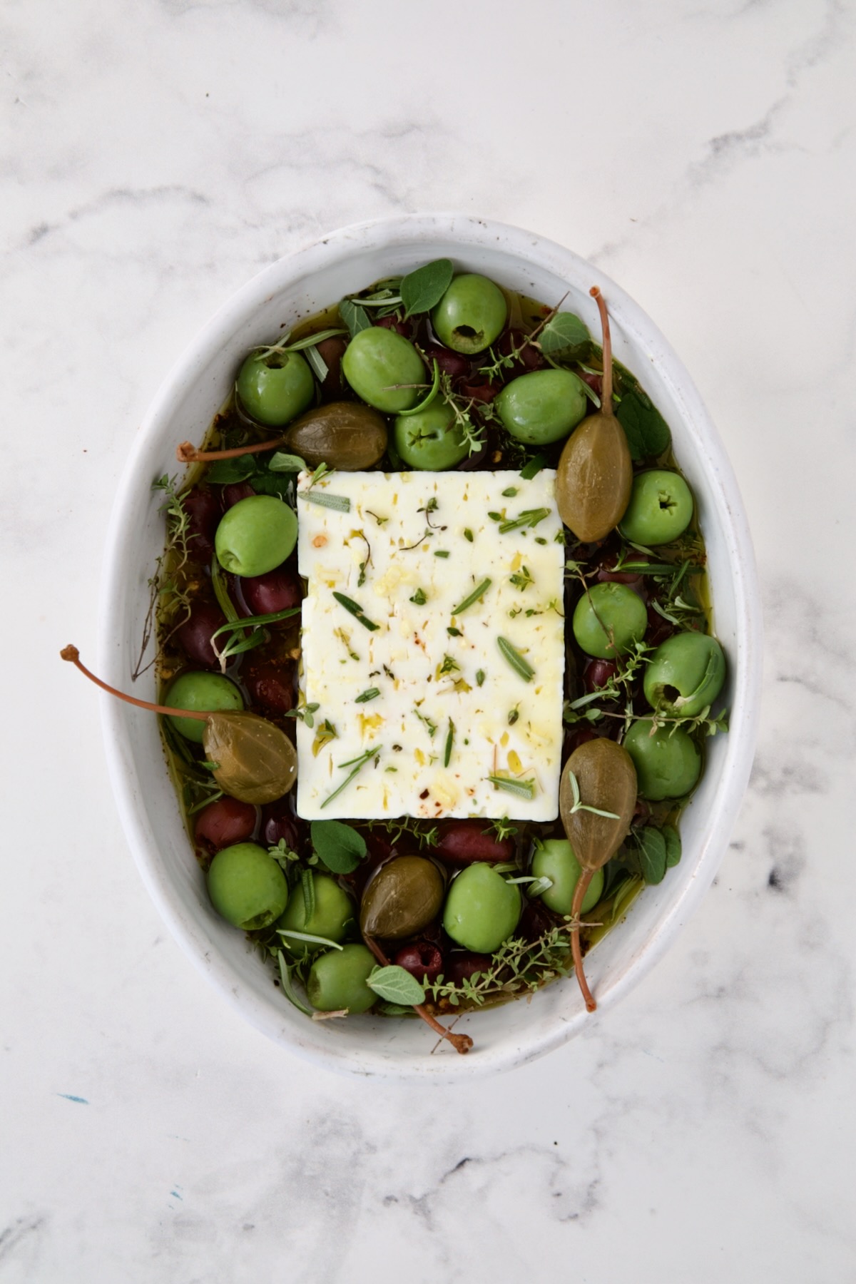 Block of feta cheese in oval white baking dish with olive oil poured over it and garlic, crushed red pepper flakes, sun-dried tomatoes, olives and caper berries added.