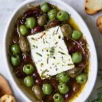 Baked Feta Cheese and Olives in white oval baking dish with serving knife surrounded by grilled bread slices.