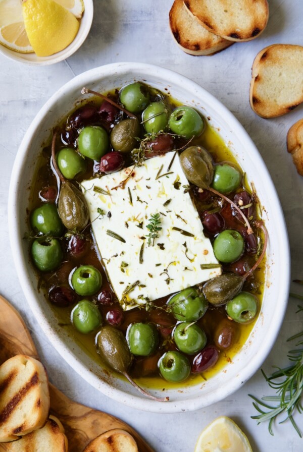 Baked Feta Cheese and Olives in white oval baking dish with serving knife surrounded by grilled bread slices.