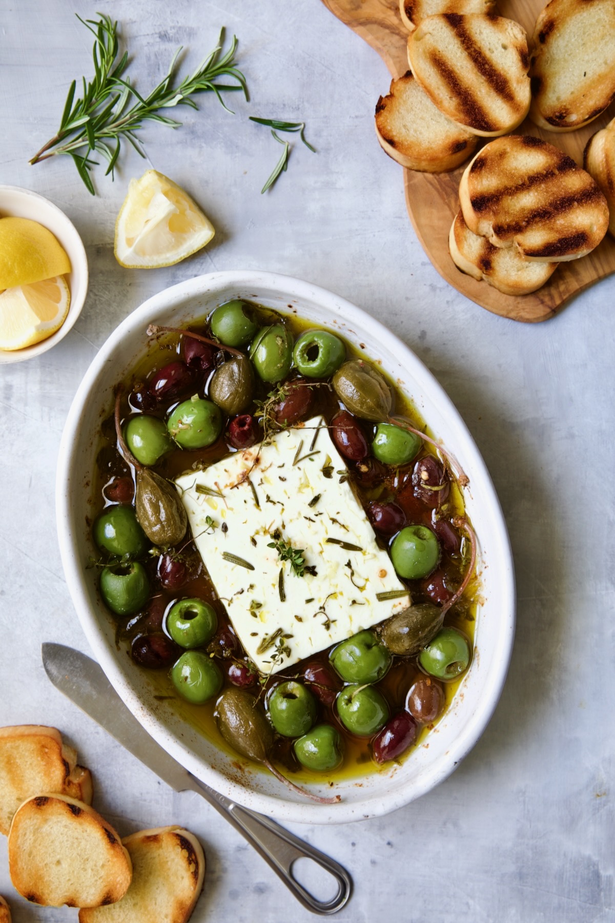 Baked Feta Cheese and Olives in white oval baking dish with serving knife surrounded by grilled bread slices and lemon wedges.