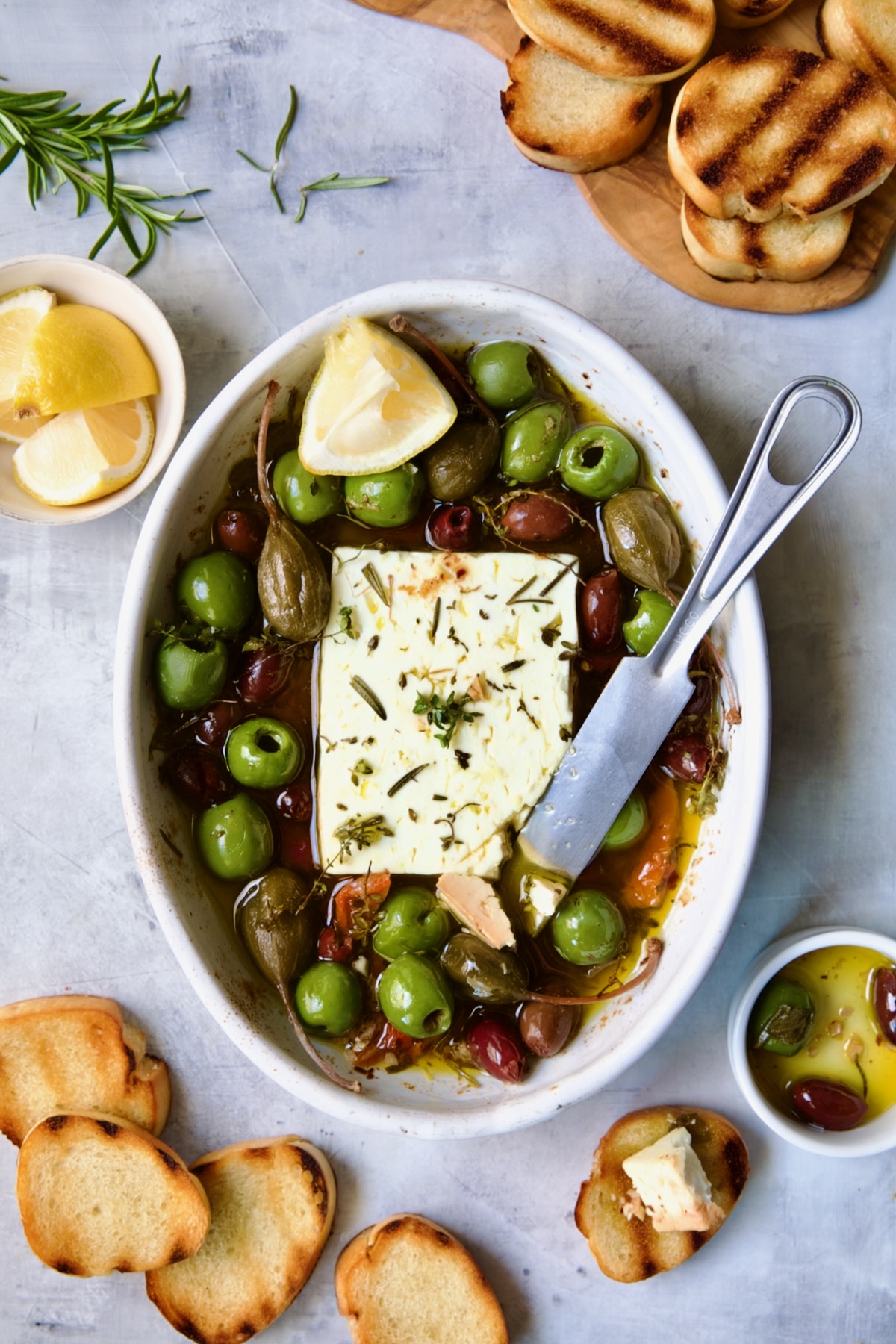 Baked Feta Cheese and Olives in white oval baking dish with serving knife surrounded by grilled bread slices.