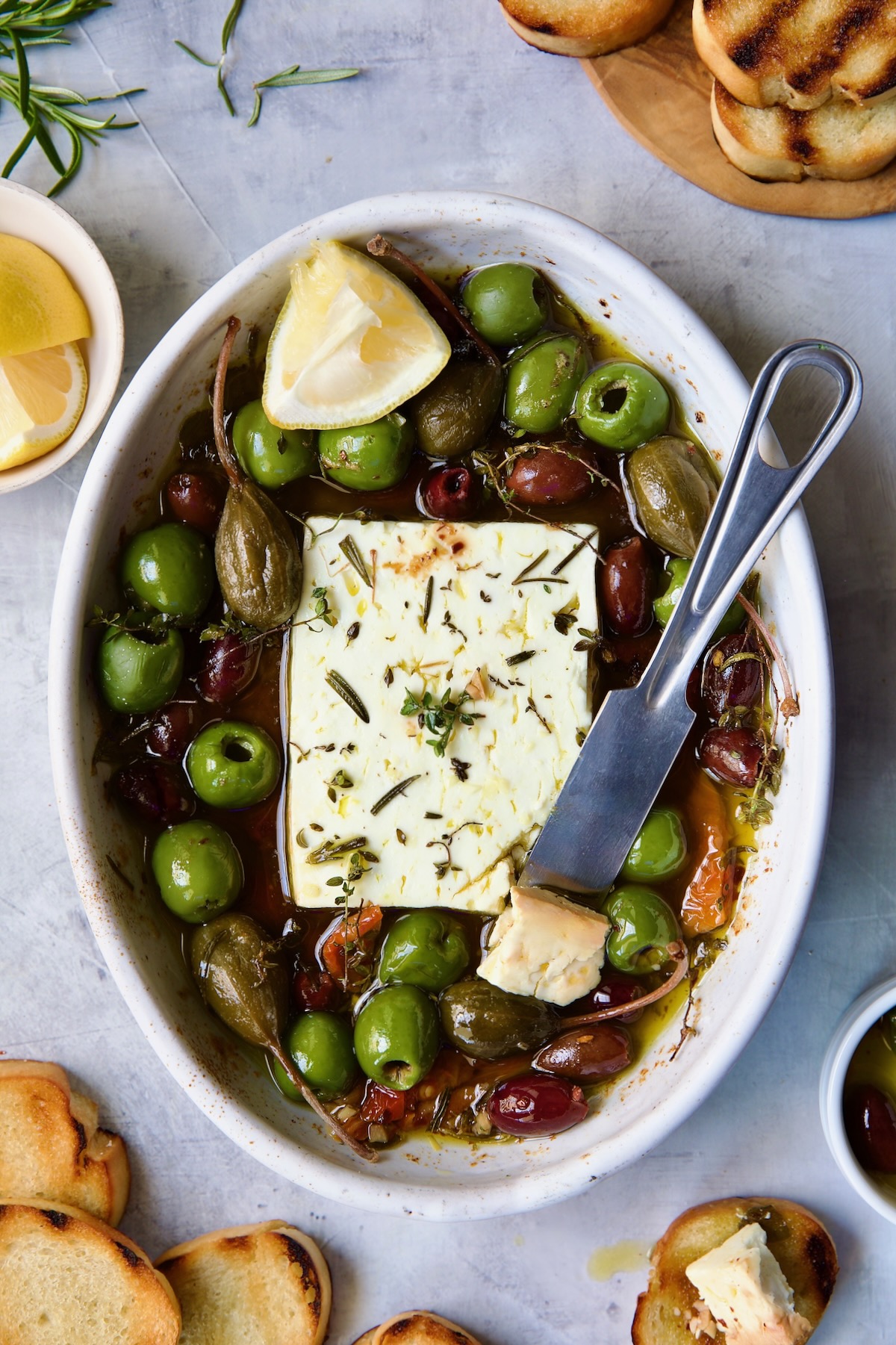 Baked Feta Cheese and Olives in white oval baking dish with serving knife surrounded by grilled bread slices.