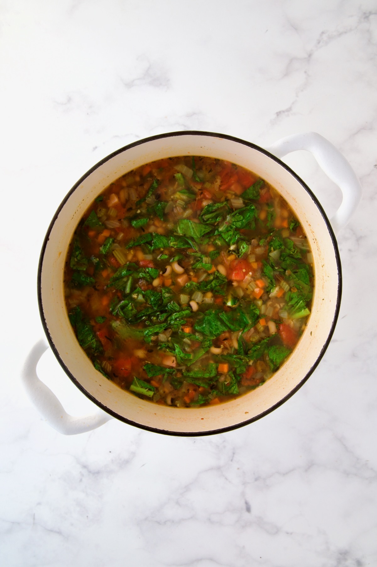 Greens and black-eyed peas added to cooked vegetables, broth, tomatoes and spices in white Dutch oven.