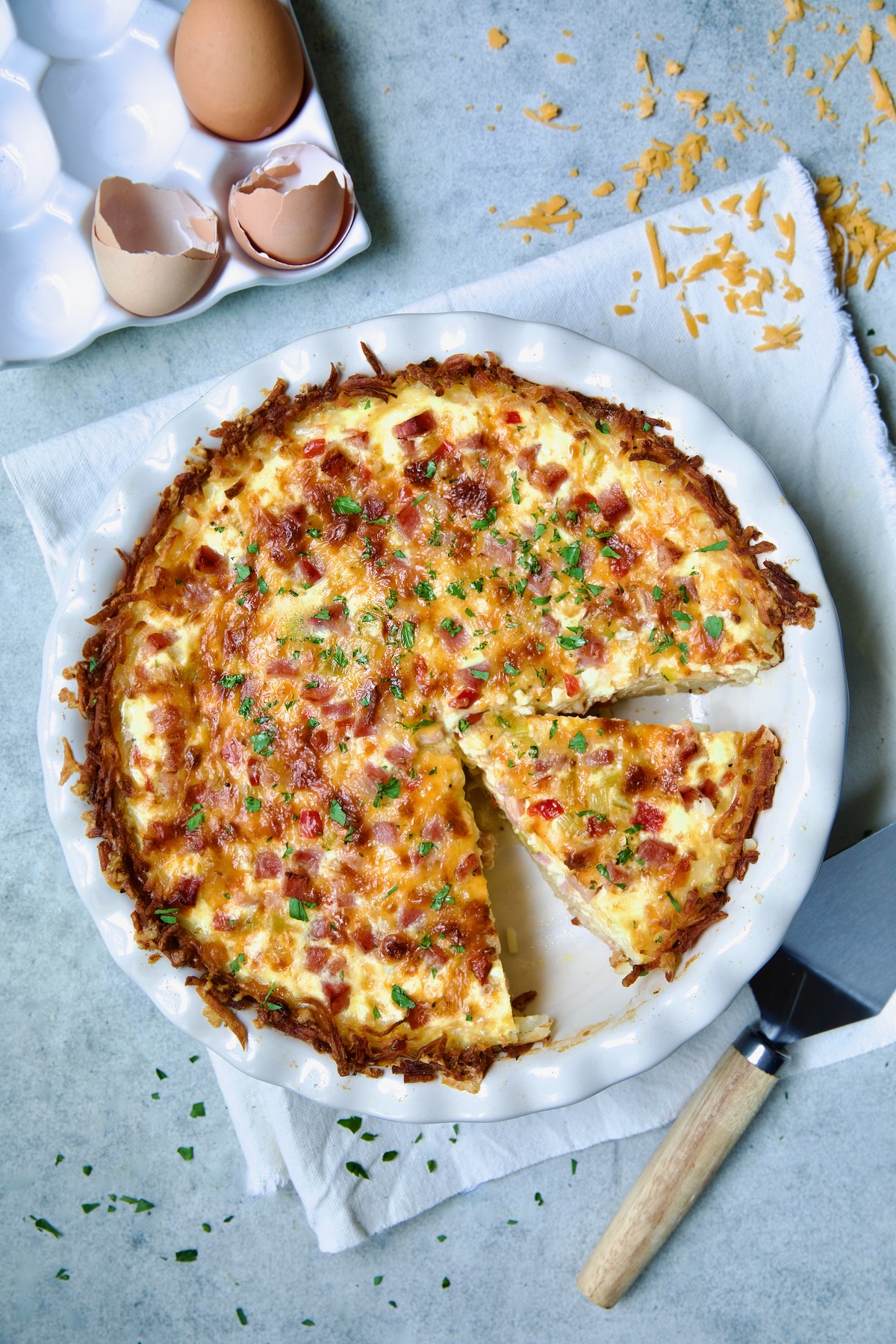 Quiche with Hash Brown Potato Crust in white pie pan on folded frayed towel with a wedge cut.