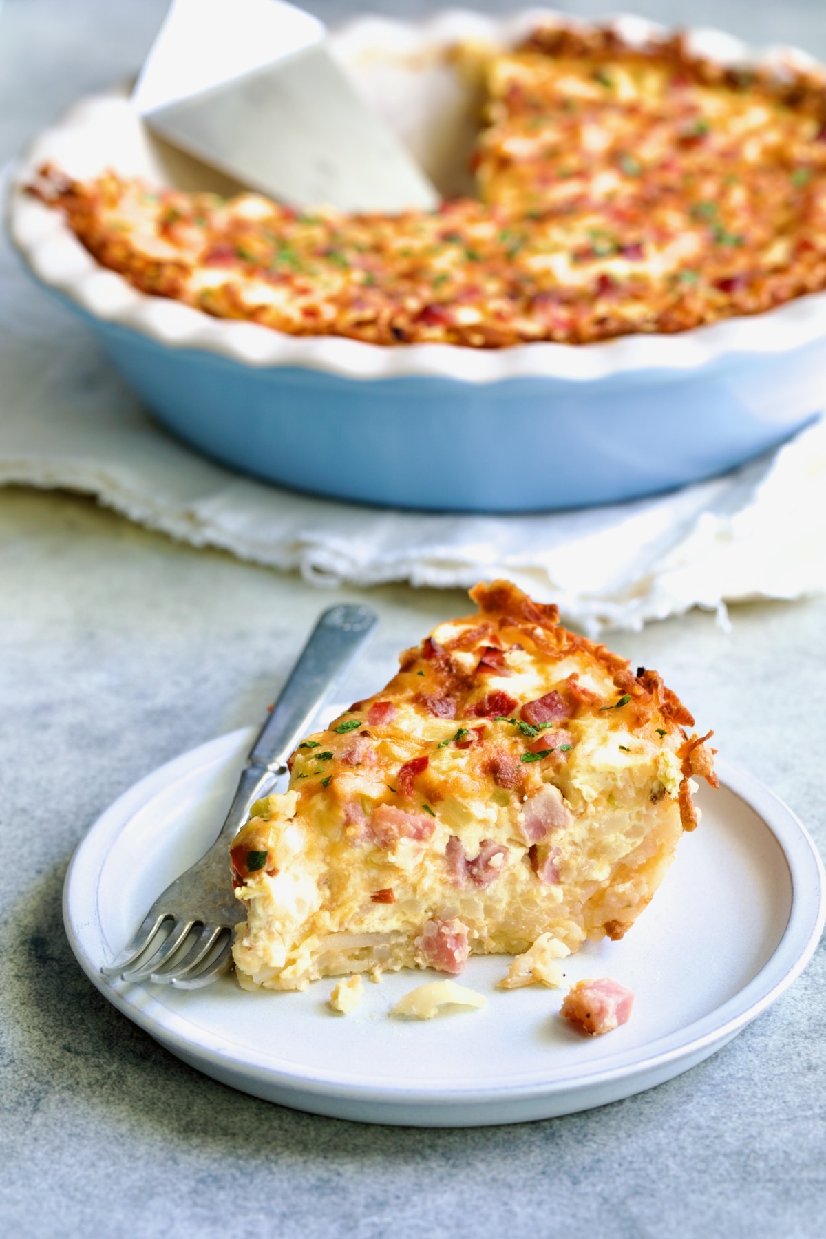 Piece of Quiche with Hash Brown Potato Crust on small white plate with fork with remaining quiche in the background.