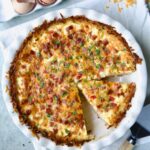Quiche with Hash Brown Potato Crust in white pie pan on folded frayed towel with a wedge cut.