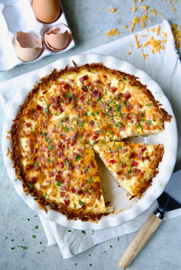 Quiche with Hash Brown Potato Crust in white pie pan on folded frayed towel with a wedge cut.