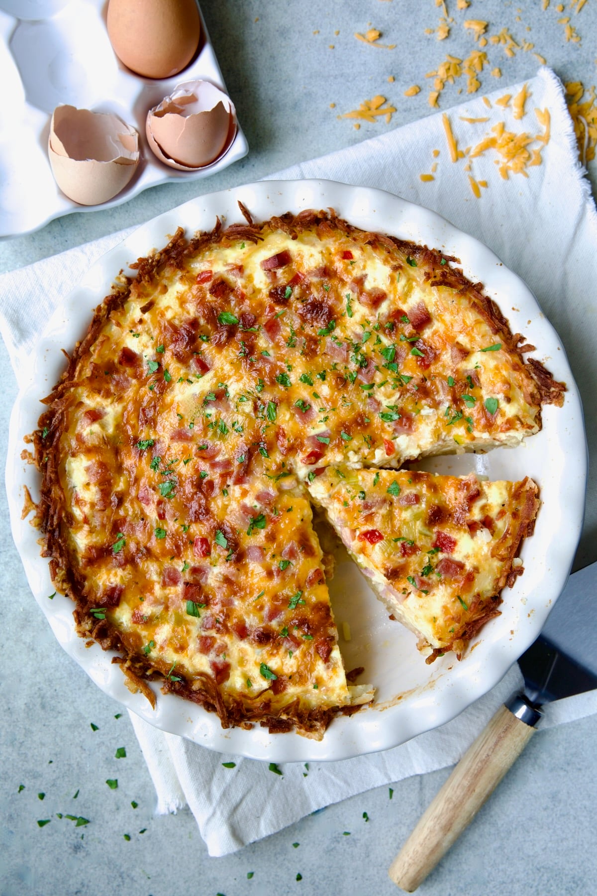 Quiche with Hash Brown Potato Crust in white pie pan on folded frayed towel with a wedge cut.