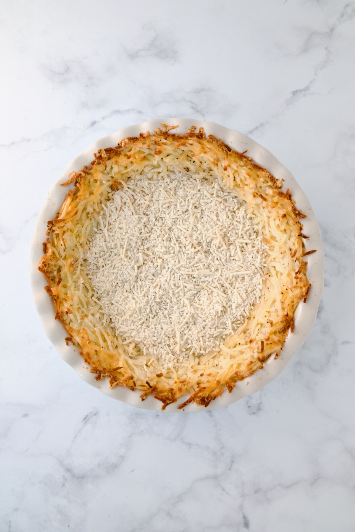 Baked hash brown potato crust in white pie pan with flour and Parmesan cheese sprinkled over the bottom.