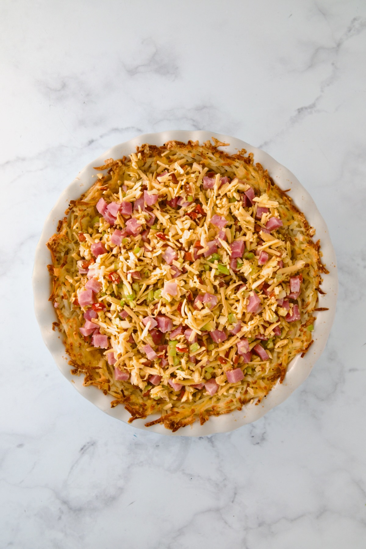 Cheese, ham and vegetables added to baked hash brown potato crust in white pie pan.