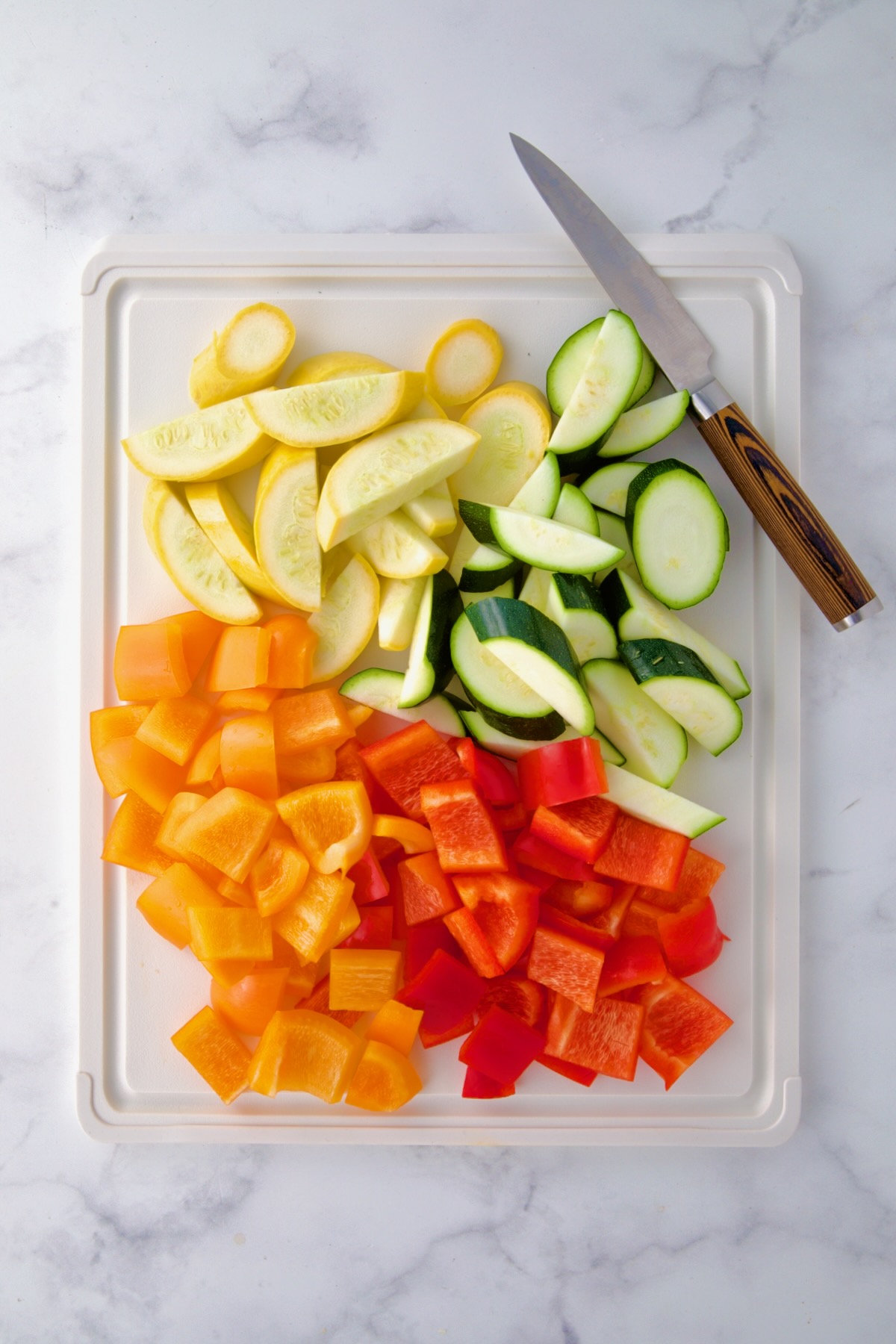 Cut yellow squash, zucchini and sweet bell peppers on white cutting board with knife.