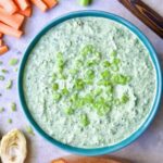 Spinach Artichoke Dip in round blue bowl garnished with scallions and carrot and celery sticks and bread round the bowl.