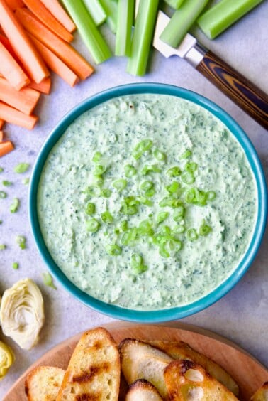 Spinach Artichoke Dip in round blue bowl garnished with scallions and carrot and celery sticks and bread round the bowl.