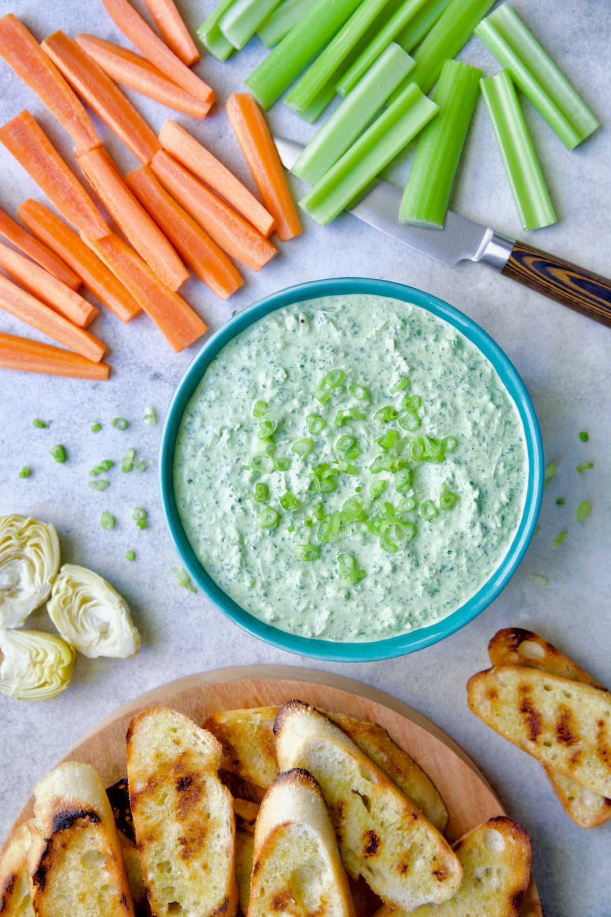 Spinach Artichoke Dip in round blue bowl garnished with scallions and carrot and celery sticks and bread round the bowl.