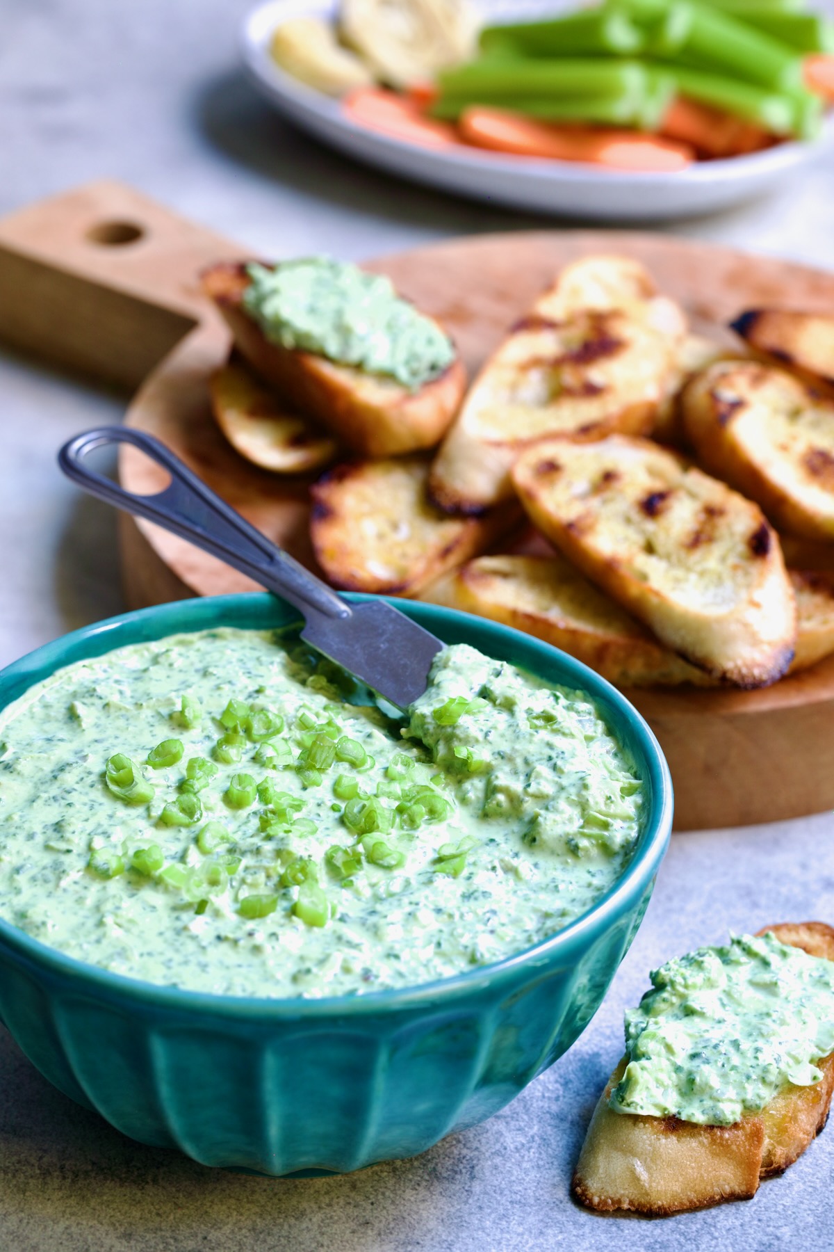 Spinach Artichoke Dip in round blue bowl garnished with scallions and carrot and celery sticks and bread round the bowl.