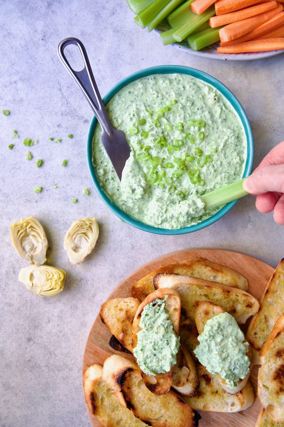 Spinach Artichoke Dip in round blue bowl garnished with scallions and carrot and celery sticks and bread round the bowl and person dipping celery stick into dip,.