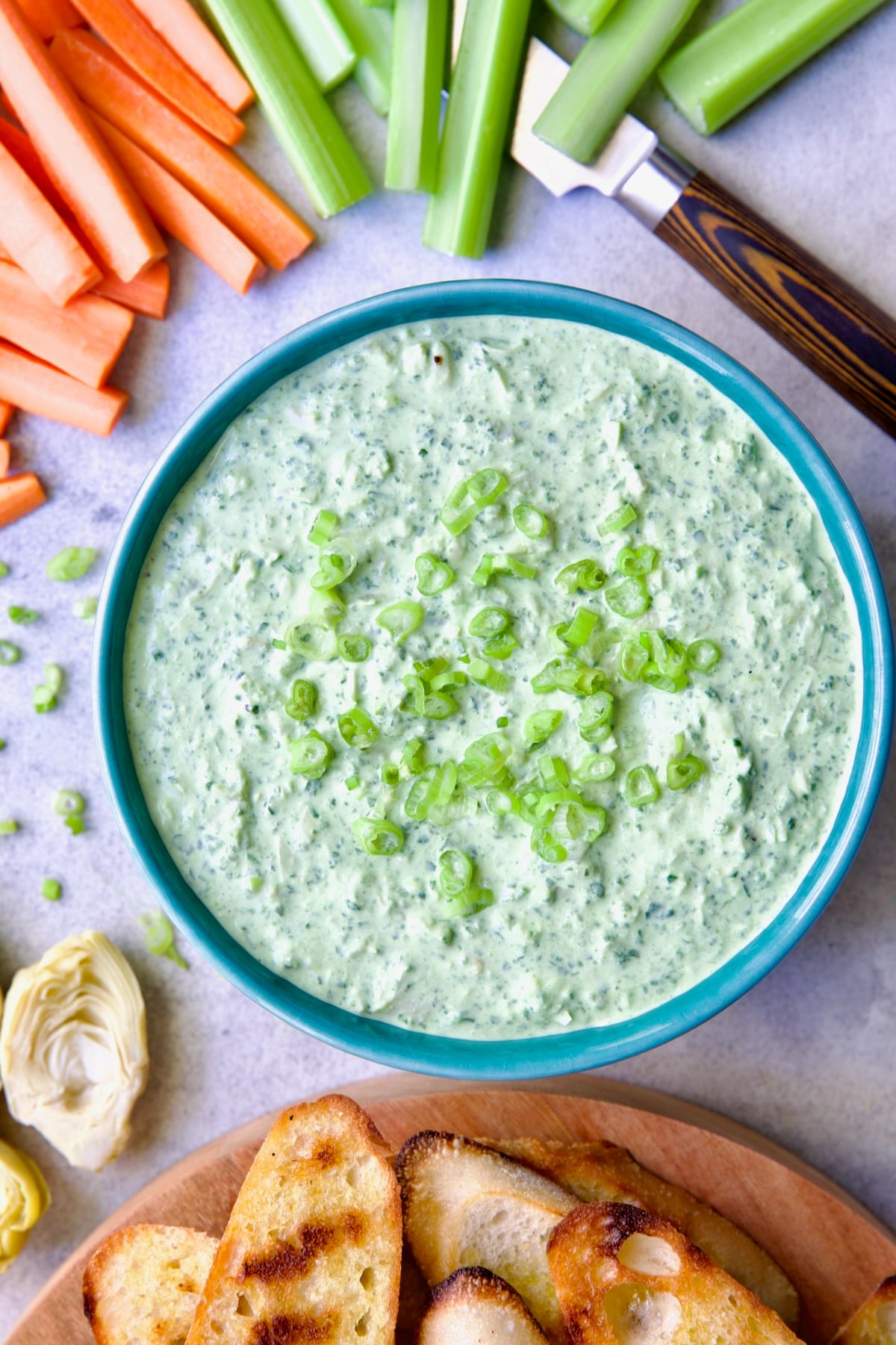 Spinach Artichoke Dip in round blue bowl garnished with scallions and carrot and celery sticks and bread round the bowl.