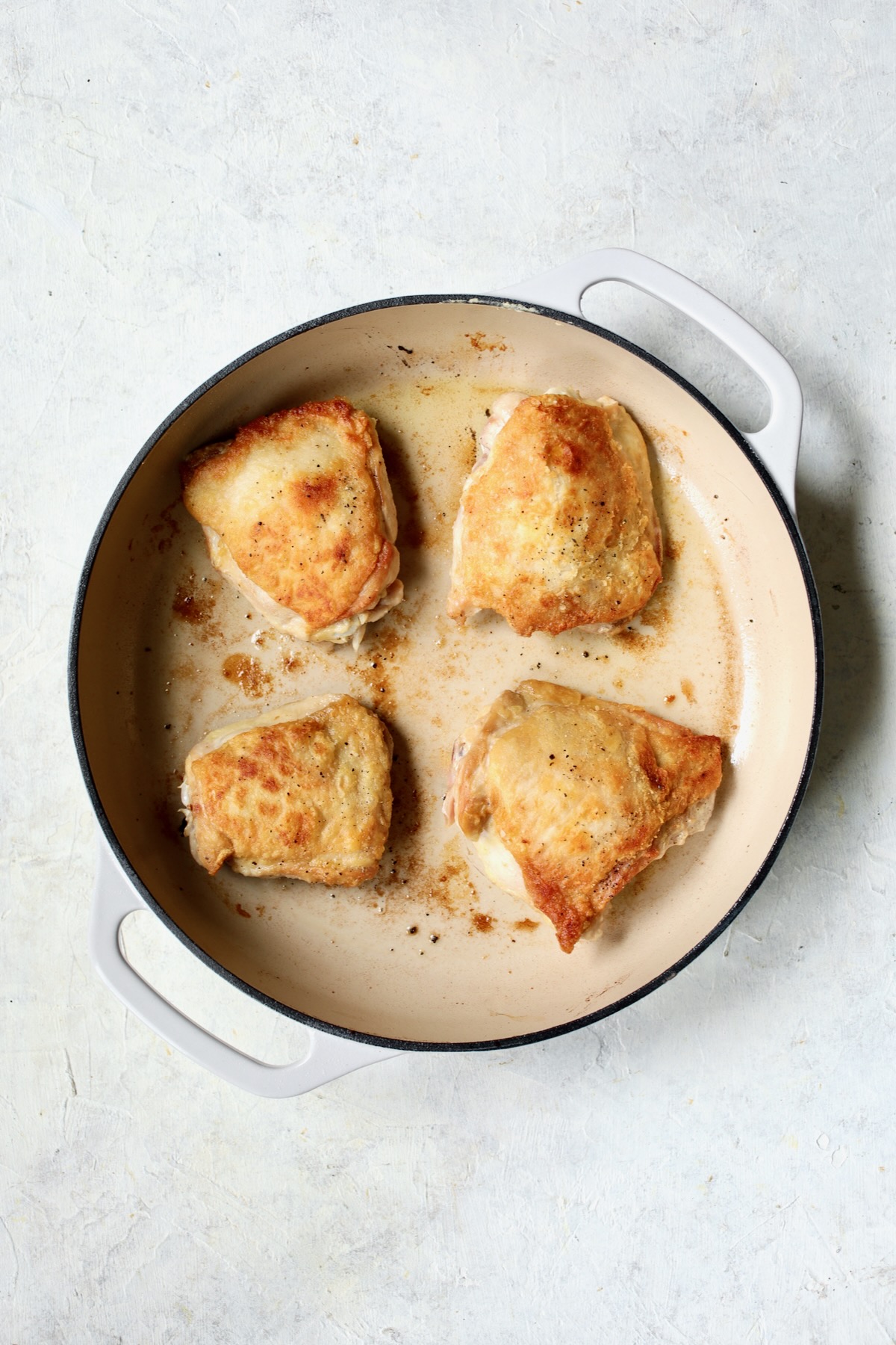 Four browned chicken thighs skin side up in white enamel cast iron skillet.