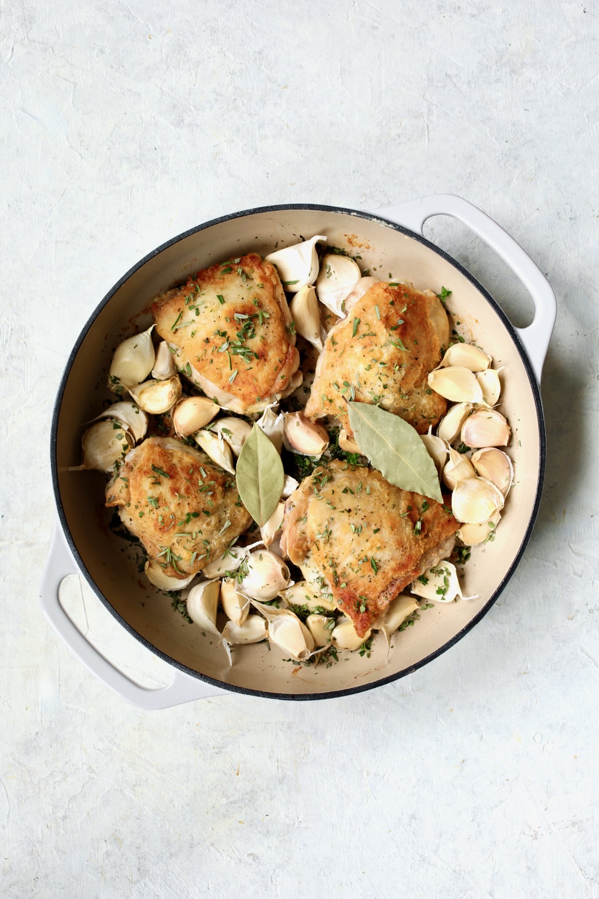 Unpeeled garlic cloves added to browned chicken thighs in white enamel cast iron skillet.