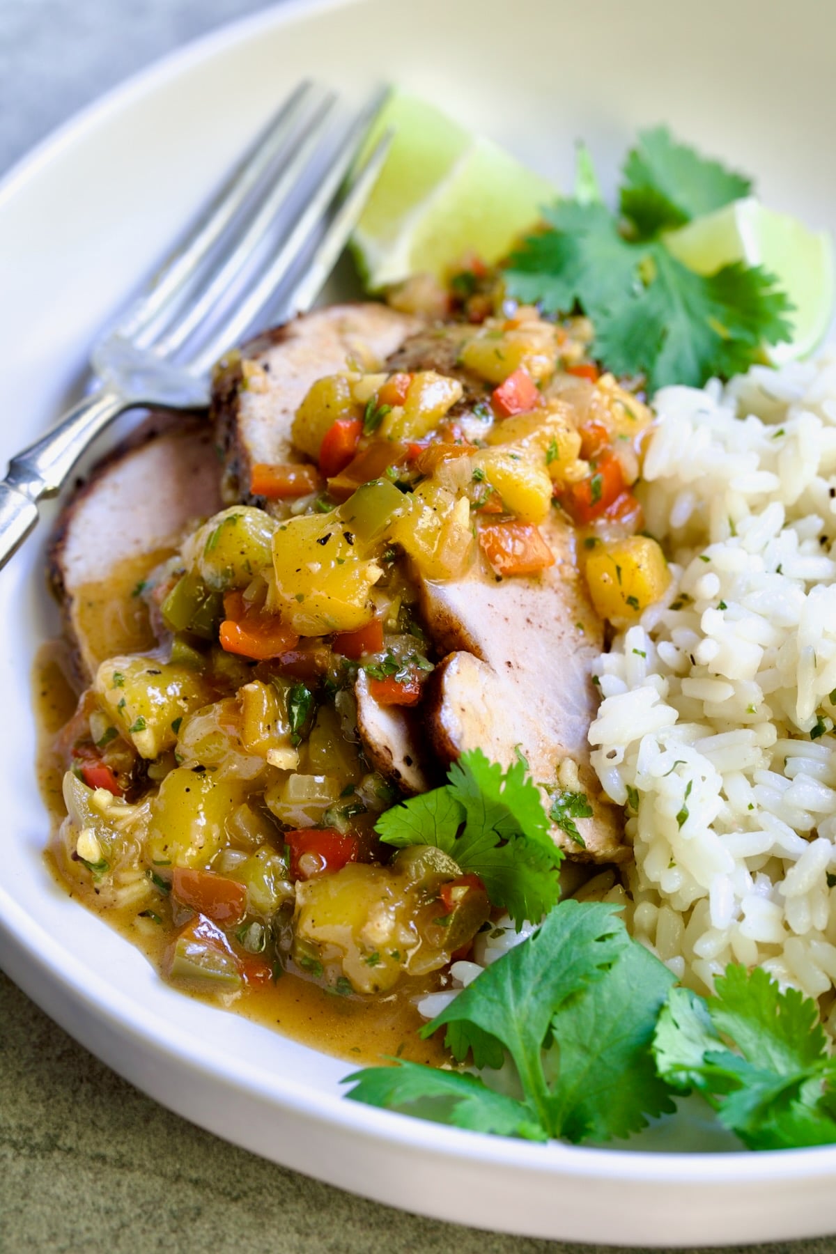 One serving of sliced and sauced Caribbean Pork Tenderloin in white dinner bowl garnished with cilantro and lime wedges.