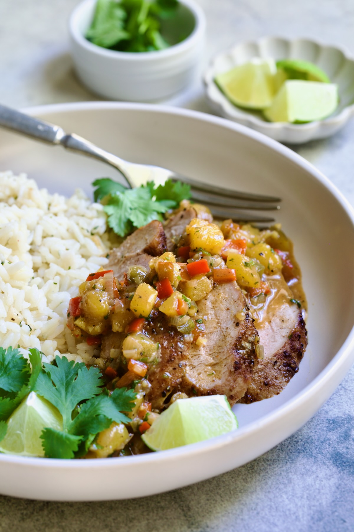 One serving of sliced and sauced Caribbean Pork Tenderloin in white dinner bowl garnished with cilantro and lime wedges.