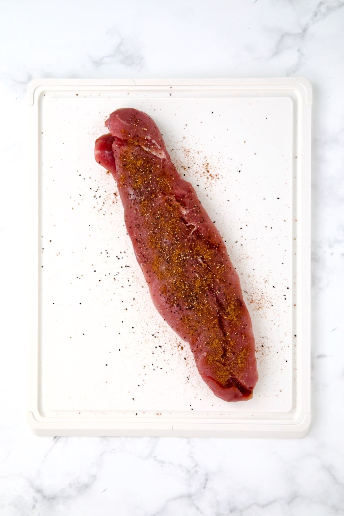 Seasoned pork tenderloin on white plastic cutting board.