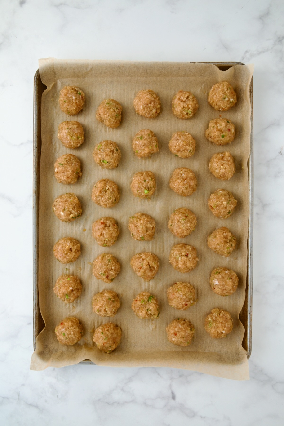 Uncooked chicken meatballs on parchment paper-lined rimmed baking sheet.