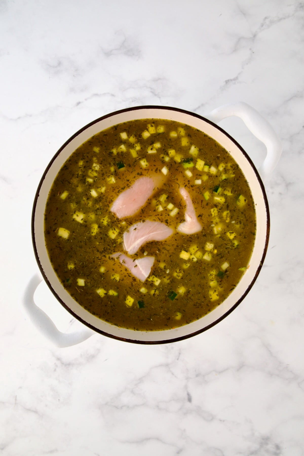 Raw chicken added to broth and vegetables in white Dutch oven.