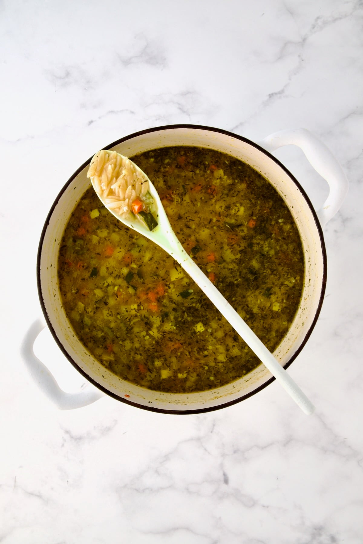 White mixing spoon full of orzo resting on white Dutch oven over soup.
