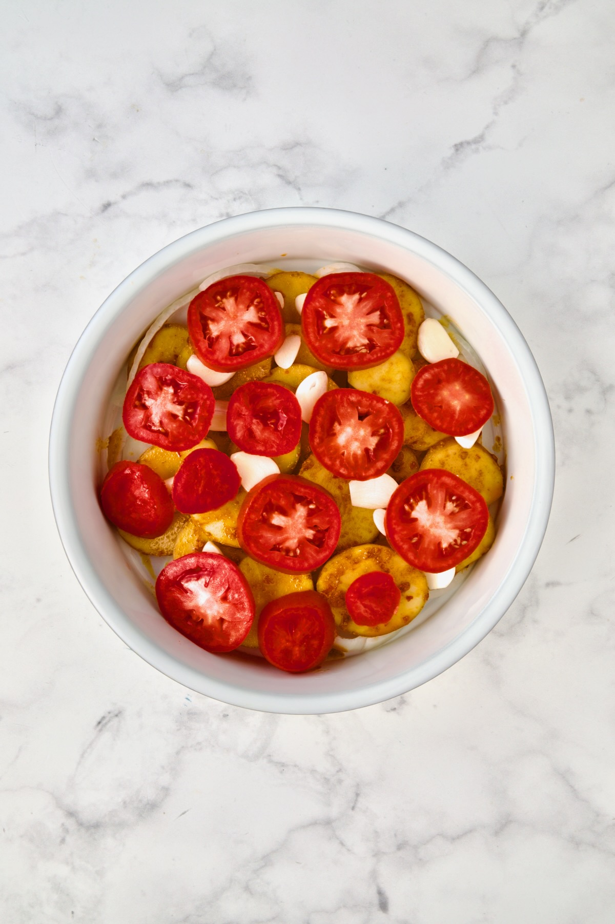 Tomatoes placed on top of potatoes, garlic and onions in round white baking dish.