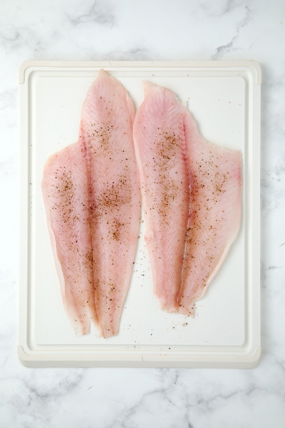 Two uncooked flounder fillets on white plastic cutting board.
