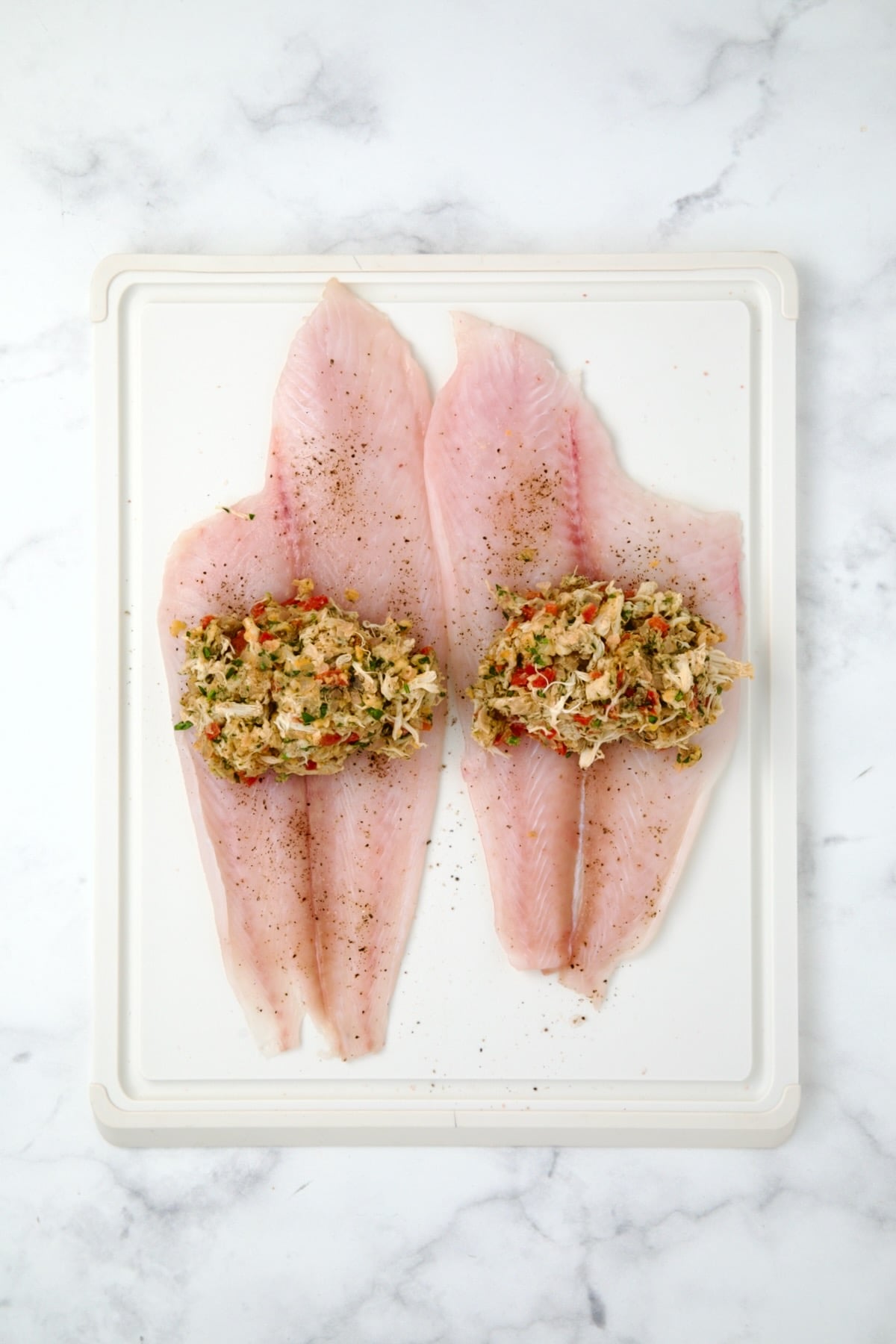 Two uncooked flounder fillets on white plastic cutting board with crab filling added.