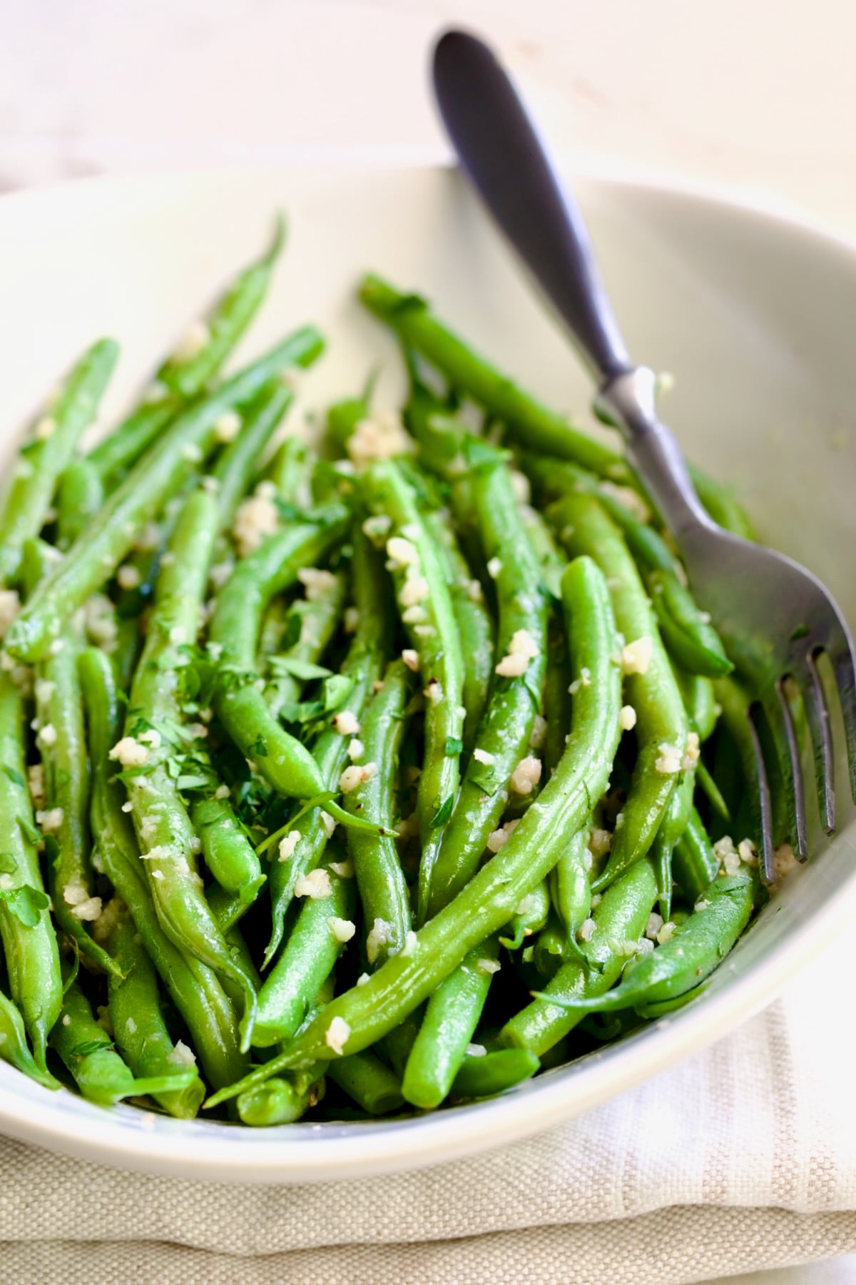 Garlic Green Beans in oval white serving bowl on folded beige towel with serving fork.