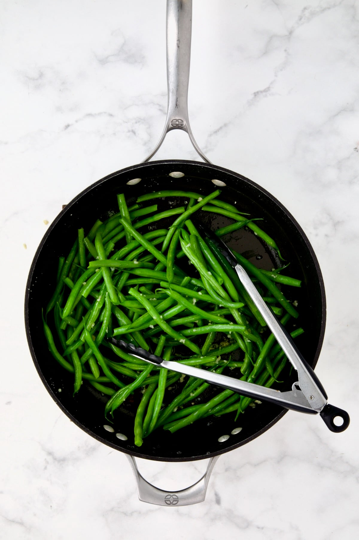 Green beans being tossed with sauteed garlic in nonstick skillet with stainless steel tongs.