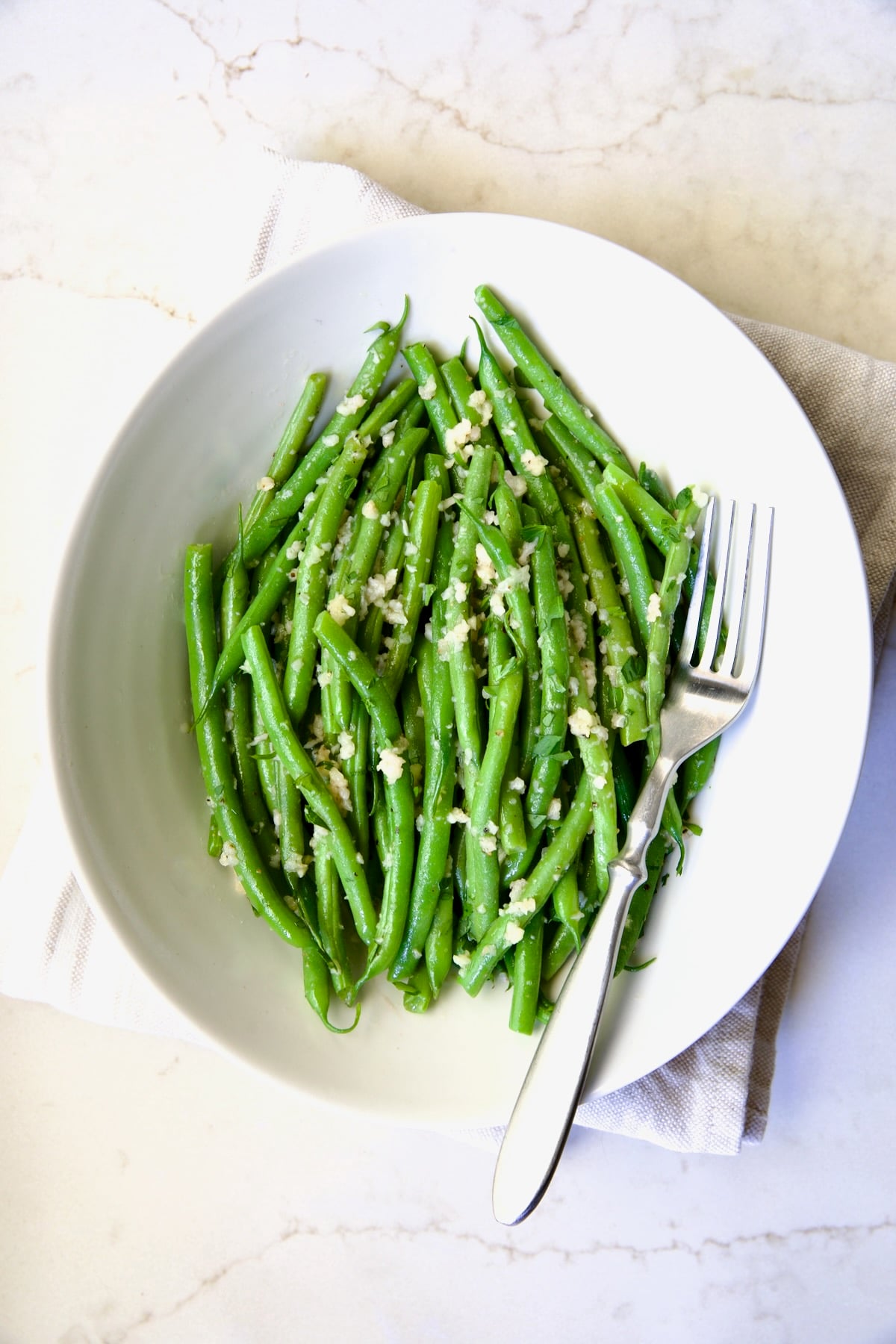 Garlic Green Beans in oval white serving bowl on folded beige towel with serving fork.