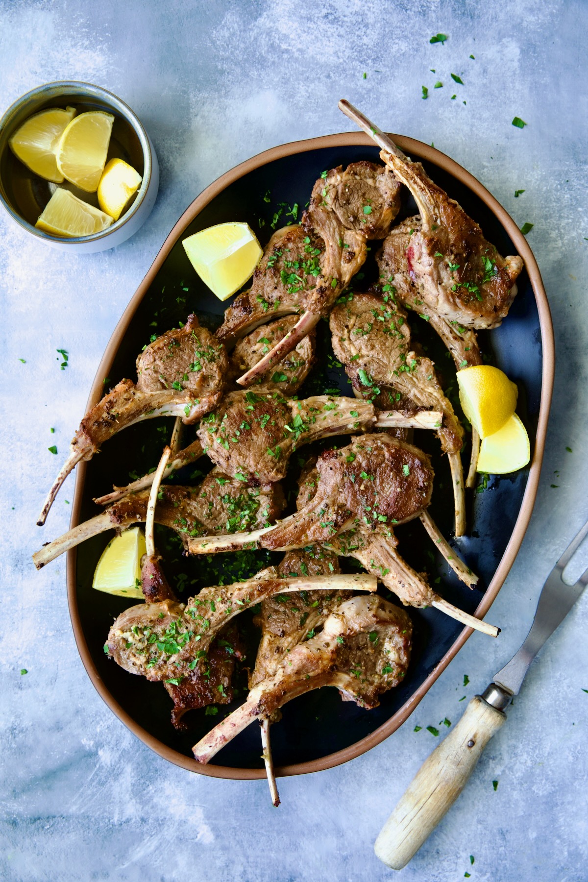 Greek Lamb Chops on black oval platter with meat fork garnished with lemon wedges.