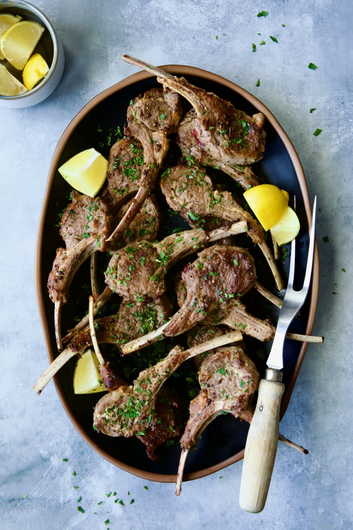 Greek Lamb Chops on black oval platter with meat fork garnished with lemon wedges.