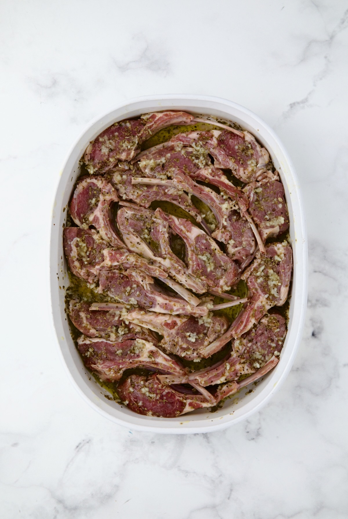 Lamb rib chops in marinade in white oval baking dish.