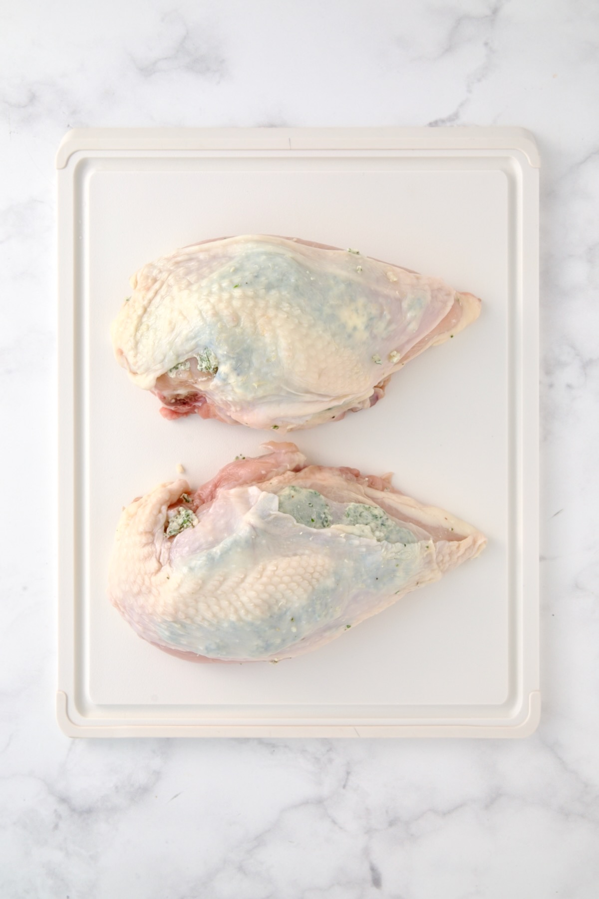 Two uncooked split chicken breasts on white plastic cutting board.