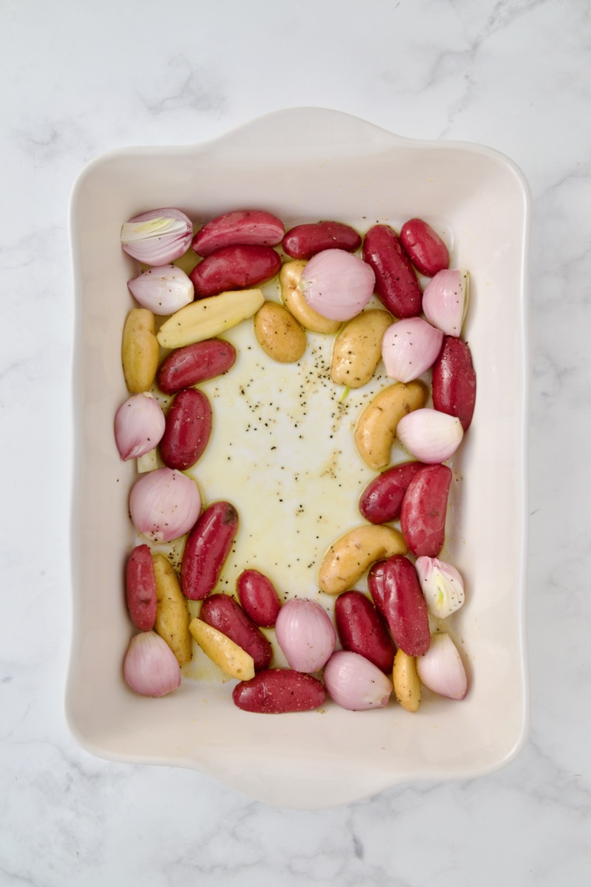 Uncooked potatoes and shallots in white rectangular baking dish.