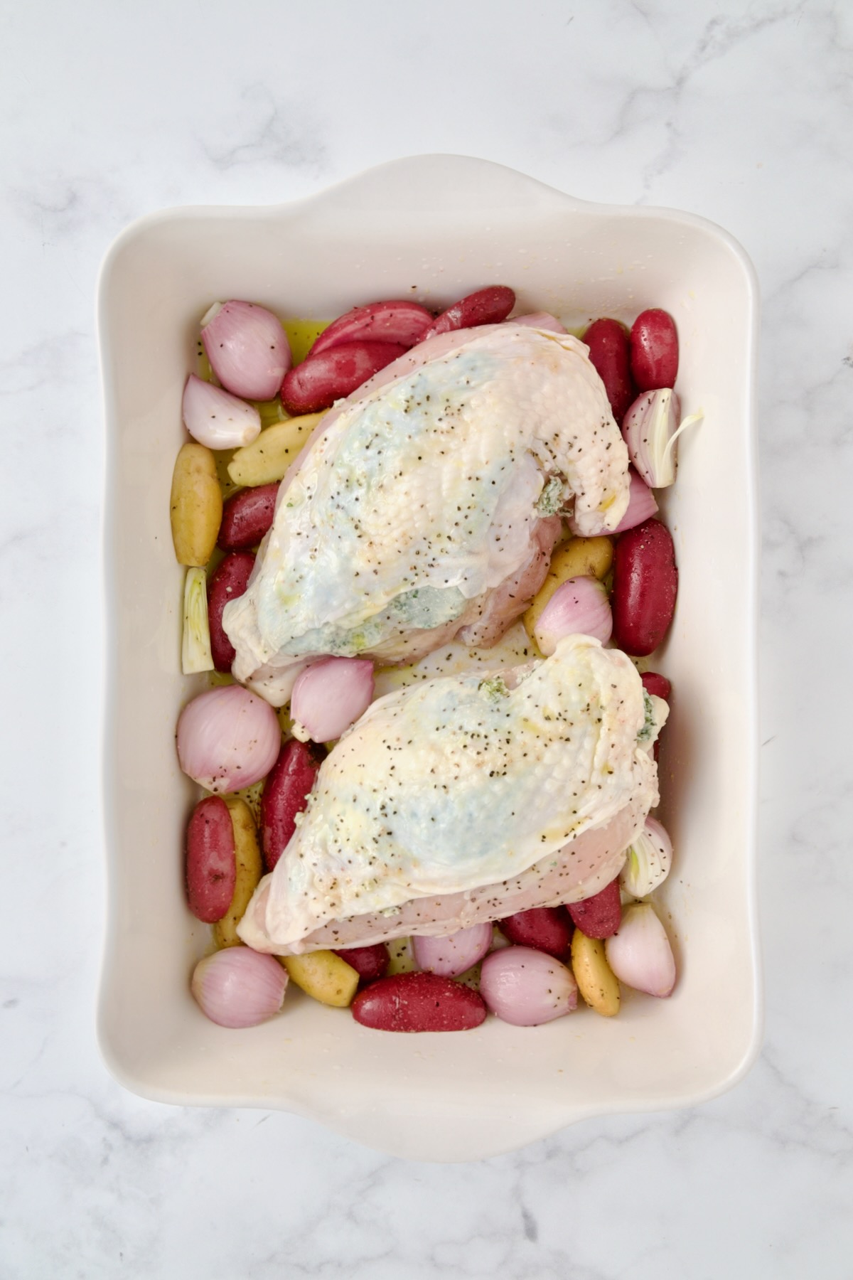 Uncooked potatoes and shallots in white rectangular baking dish with uncooked split chicken breasts placed on top.